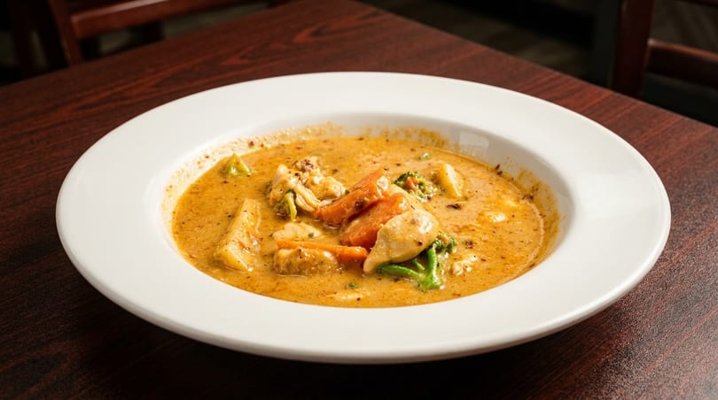 Creamy yellow curry with chicken, carrots, and broccoli in a white bowl on a wooden table.